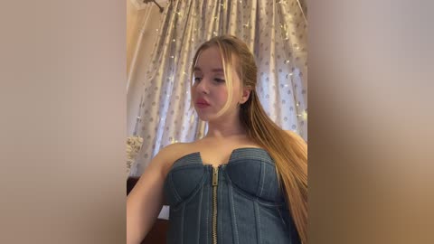 A video of a young Caucasian woman with long, straight blonde hair, wearing a strapless denim corset, standing in a room with white lace curtains and fairy lights in the background.