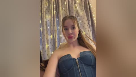 Video of a young woman with fair skin and long blonde hair, wearing a strapless denim corset, against a background of beige curtains with fairy lights.