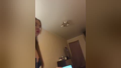Video of a young woman with long brown hair and a slim figure, standing in a dimly lit room with beige walls, a ceiling fan, and a wooden door.