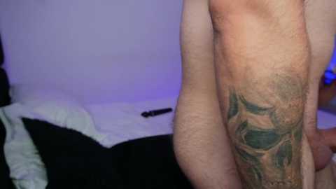 Video of a close-up of a man's leg with a detailed green tattoo of a skull and crossbones on his calf, in a dimly lit bedroom with a white bedsheet and purple lighting.