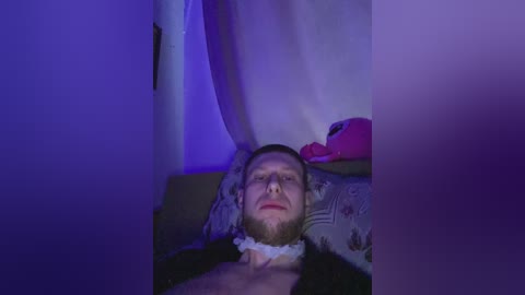 Video of a man with a red beard, lying on a bed, under purple lighting, wearing a white bandage on his head, surrounded by a dimly lit, cluttered room.