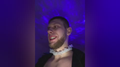 Video of a shirtless, muscular, light-skinned man with a trimmed beard, wearing a white beaded necklace, against a purple backdrop. He appears to be laughing.