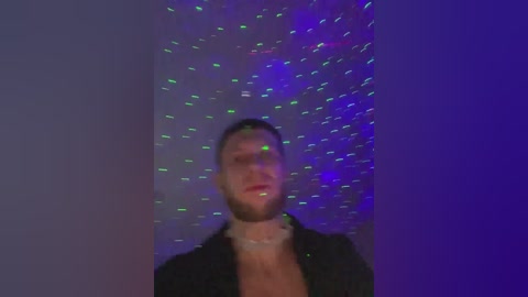 A grainy, low-light video captures a man with a trimmed beard and short hair, wearing a black shirt. The background is a dark, blurry room with colorful, neon-lit confetti floating around. The image has a vintage, slightly out-of-focus quality.