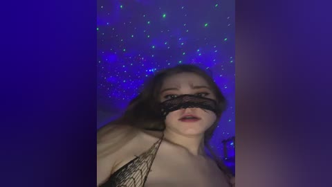 Video of a fair-skinned woman with long brown hair, wearing a black lace blindfold, standing against a purple, starry background. She is topless, with a hint of cleavage visible.