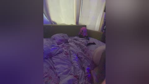 A dimly-lit video of a messy, unmade bed with colorful, patterned bedding, featuring stuffed toys, in a small, cozy room with white curtains.
