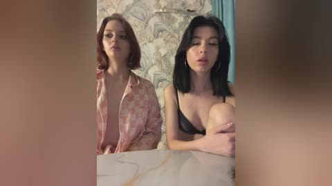 Media: Video of two women in a bathroom: one in a pink floral robe, the other in a black bra, both with fair skin, dark hair, and closed eyes, leaning against a marble countertop.