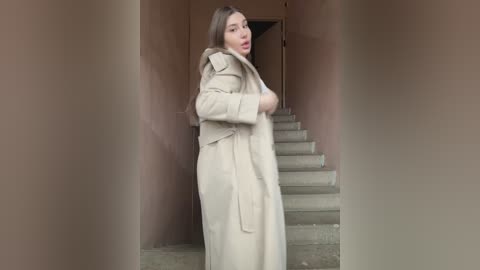 Media: A video of a fair-skinned woman with long brown hair, wearing a beige trench coat, standing on concrete steps, with a muted brown wall in the background.