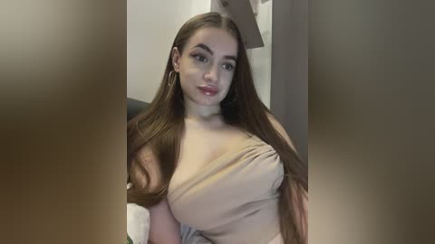 Video of a young Caucasian woman with long, straight brown hair and fair skin, wearing a beige, off-the-shoulder top, sitting indoors, blurred background.