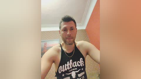 Media: Video of a muscular, light-skinned man with short dark hair and a beard, wearing a black \"Outback\" tank top, standing indoors with orange and beige walls, a ceiling light, and a framed picture in the background.