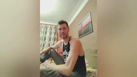 Media: A video of a man with short brown hair, a beard, and a muscular build, wearing a black tank top and camouflage pants, sitting on a bed with floral-patterned sheets. The room features beige wallpaper and a framed picture of a man on a horse.