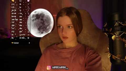 Video of a fair-skinned woman with brown hair wearing a pink shirt, seated in a plush chair with a microphone. Background features a digital star chart and dim lighting.