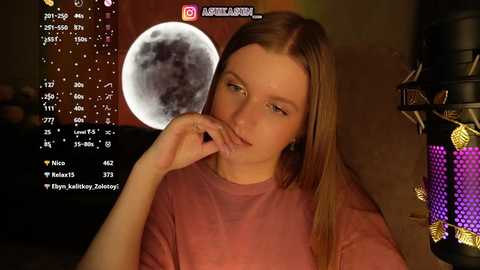 Video of a young woman with light skin and straight brown hair, wearing a pink t-shirt, looking pensively at a full moon overlay on her screen. Background includes a purple and black microphone stand.