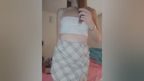 Video of a slender, fair-skinned woman with straight, shoulder-length brown hair, wearing a white crop top and high-waisted plaid skirt, posing in a dimly lit bedroom with messy bedding.