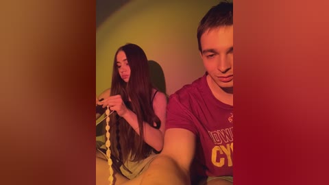 Video of two young people, a brunette girl in a green dress and a short-haired boy in a maroon shirt, sitting closely, with a warm, reddish-yellow light casting a cozy atmosphere.