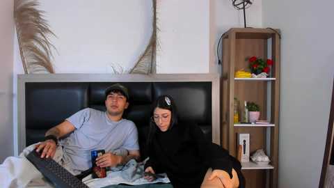 A video of a young Asian couple in a modern bedroom; he wears a gray hoodie, she a black dress, both asleep on a black headboard.