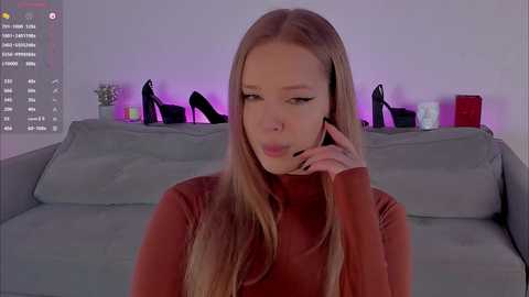 Video of a young Caucasian woman with long blonde hair, wearing a red turtleneck, talking on a phone while sitting on a gray couch. Background features purple LED lighting and black high heels.