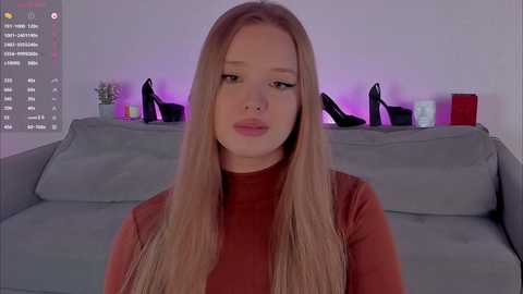 Video of a young Caucasian woman with long, straight blonde hair, wearing a red turtleneck, seated in a modern living room with a grey sofa and black high heels displayed on a shelf behind her.