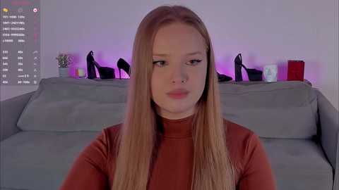 Video of a fair-skinned, blonde-haired woman with a nose ring, wearing a high-necked rust-colored top, sitting on a gray couch in a dimly lit room with purple lighting and black high heels on a shelf.