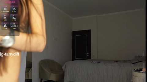 Media: Video of a dimly lit, sparsely furnished bedroom with a beige bed, a dark wooden door, and a chair. The room has a minimalist, somewhat neglected appearance.