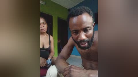 Video: Black man with short hair, shirtless, smiling, leaning forward; woman with medium skin tone, wearing a black tank top, sitting, expression neutral. Background: bright green wall, blue wall, doorway, dim lighting.