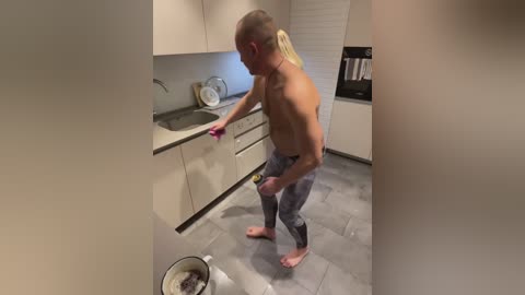 Media: Video of a shirtless, muscular man with a ponytail, wearing grey pants, washing dishes in a modern kitchen with white cabinets and a grey countertop.
