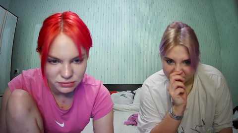 Media: Video of two women with bright red and blonde hair, wearing casual pink and white t-shirts, respectively, sitting on a bed with white sheets in a simple, green-patterned wallpapered room.