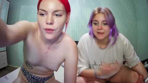 Media: Video of two young women with pale skin, one with red hair, the other with lavender hair, both topless, wearing leopard-print underwear, sitting on a bed in a dimly lit room with green walls.