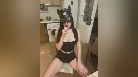 A video of a fair-skinned woman with long black hair, wearing a black cat mask and black lingerie, seated in a modern kitchen with wooden cabinets and white appliances.