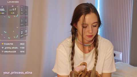 Video of a young woman with long brown hair in braids, wearing a white t-shirt with a cartoon design, in a room with vertical blinds and a bed.