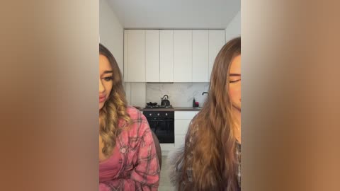 Media: Video of two women with long, wavy hair, one in pink plaid pajamas, the other in a gray hoodie, standing side-by-side in a modern kitchen with white cabinets and marble backsplash.
