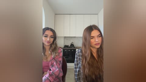 Media: Video of two young women, one with long, wavy brown hair, the other with straight, light brown hair, standing in a modern kitchen with white cabinets, black stove, and marble backsplash.