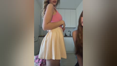 Media: Video of a slender young woman with long brown hair, wearing a pink tank top and a white pleated skirt, standing in a modern kitchen with white cabinets.