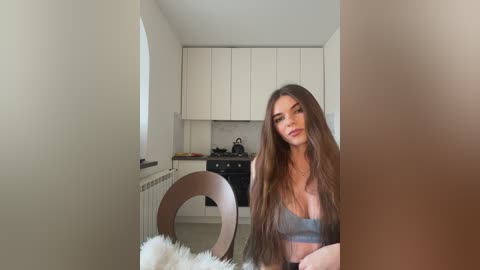 Media: Video of a woman with long brown hair, wearing a gray bralette, sitting in a modern kitchen with white cabinets and a black stove.