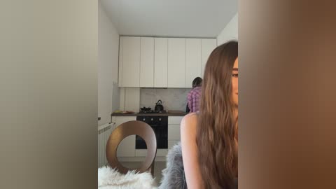 Media: Video of a modern, minimalist kitchen with white cabinets and a small, dark-haired woman in a purple sweater standing at the stove.
