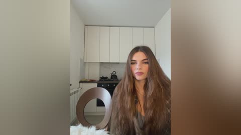 Media: A video of a young woman with long, wavy brown hair, wearing a black top, sitting at a wooden table in a minimalist kitchen with white cabinets and a stove.