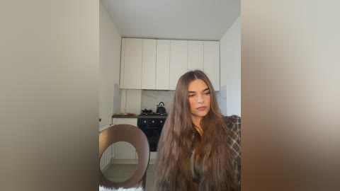 Media: Video of a young woman with long, straight brown hair, sitting at a modern kitchen table. She wears a plaid shirt. Background shows a sleek, white kitchen with stainless steel appliances.