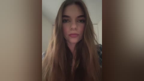 Media: A video of a young woman with long, wavy brown hair, light skin, and pink lipstick, wearing a patterned shirt, standing indoors with blurred background.