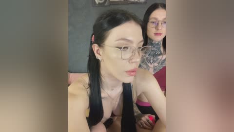 Video of two women: a slender, fair-skinned woman with black pigtails and glasses, and a tattooed, dark-haired woman with glasses. Both wear red lipstick.