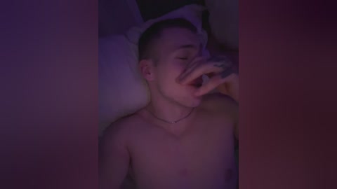 Media: Video of a young, shirtless, fair-skinned man with short hair, lying on a bed, mouth open, hand on cheek, under dim purple lighting, pillow nearby.