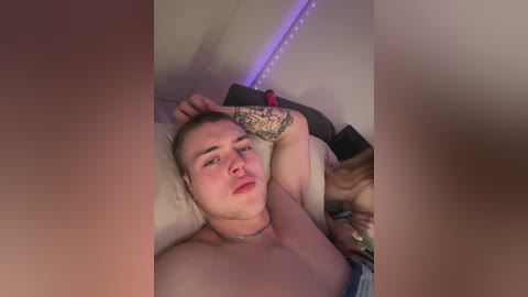 Media: Video of a shirtless, muscular young man with a tattoo on his arm, lying on a bed with a pillow, surrounded by soft lighting and a purple LED strip.