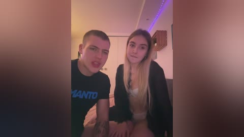 Media: Video of two young, light-skinned, slender individuals with pale skin tones, one with a shaved head, the other with long blonde hair. They are dressed casually, sitting on a bed, under soft lighting.