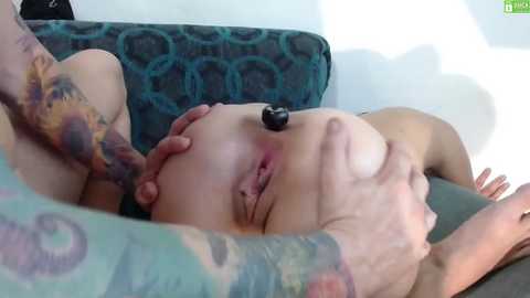 Video of a woman with tattoos, lying on a teal-patterned couch, receiving nipple play from a tattooed man with a black nipple clamp.