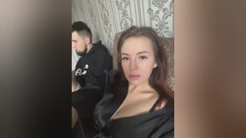 Media: A video of a man and woman in a dimly lit room. The man, with short dark hair and a beard, wears a black hoodie. The woman, with long brown hair, is in a black satin robe, revealing cleavage.