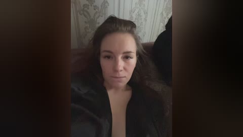 A video of a fair-skinned woman with long, brown hair, wearing a black leather jacket, lying on a dark couch against a floral-patterned wallpaper. Her expression is neutral.