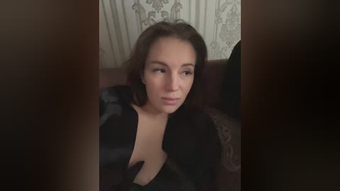 Video of a fair-skinned woman with shoulder-length brown hair, wearing a black satin robe, seated on a maroon sofa, partially obscured by a curtain.