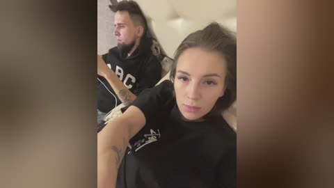 Media: Video of a bearded man with long hair and a woman with long hair, both wearing black hoodies, seated on a beige couch in a dimly lit room.