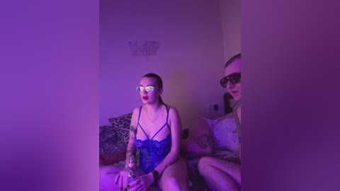 Video of a light-skinned woman with a tattooed arm, wearing a black lace bra and holding a glass dildo, sitting on a bed with a man in sunglasses, both under purple lighting.