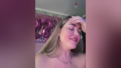 Video of a smiling, fair-skinned woman with long blonde hair, wearing a silver necklace, touching her face, in a room with pink and metallic decorations.