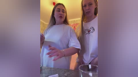 Video of two young women in white T-shirts, one with long blonde hair and the other with long brown hair, standing in a bathroom with a pink shower curtain.