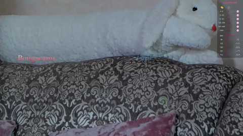Video of a plush, white, teddy bear with black eyes and a red nose lying on a bed with a black and white damask-patterned comforter. The background is a plain, light-colored wall.
